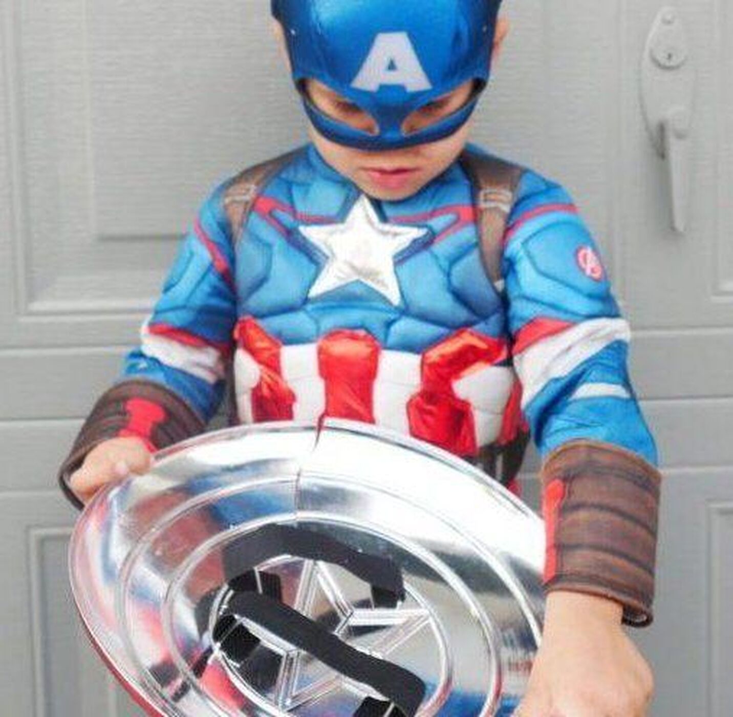 A young child dressed in a Captain America costume, including a blue mask with a white "A" and a star on the chest, stands in front of a gray garage door while looking down at a shiny toy shield held in their hands.