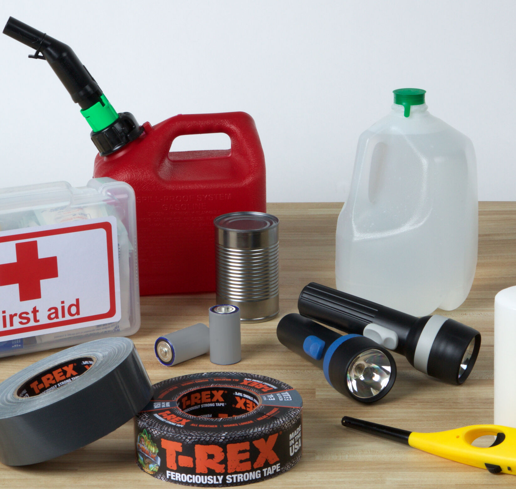 Some Emergency Preparedness Supplies on a table (First Aid Kit, Gas Can, Water Jug, Candle, Lighter, Flashlights, Batteries, Can of food, and T-Rex Duct Tape)
