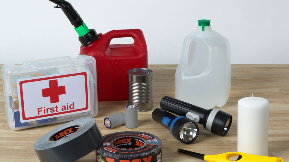 Some Emergency Preparedness Supplies on a table (First Aid Kit, Gas Can, Water Jug, Candle, Lighter, Flashlights, Batteries, Can of food, and T-Rex Duct Tape)